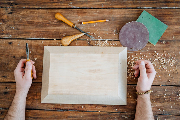 A person processes a rectangular wooden surface, sawdust is lying around and woodworking tools lie, the frame is shot from above. Cutting out parts, dishes, toys from wood. High quality photo - Foto, afbeelding