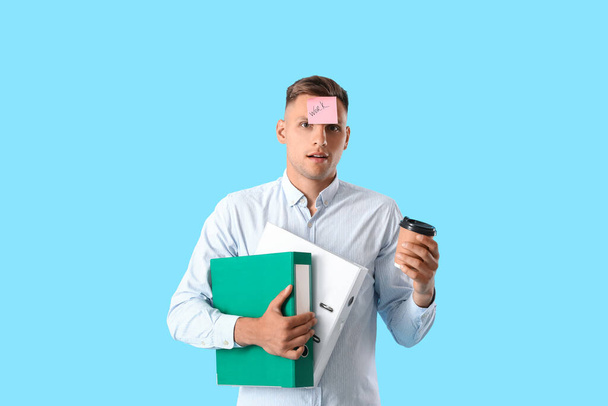Jongeman met mappen, plakkerige noot met woord WORK op zijn voorhoofd en koffie tegen kleur achtergrond - Foto, afbeelding