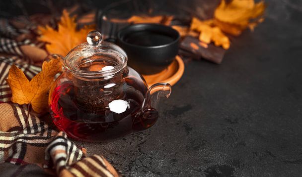 Composition d'automne sur fond sombre. Une tasse de thé et théière, écharpe, barre de chocolat et feuilles d'automne. Pose plate, vue du dessus, espace de copie - Photo, image