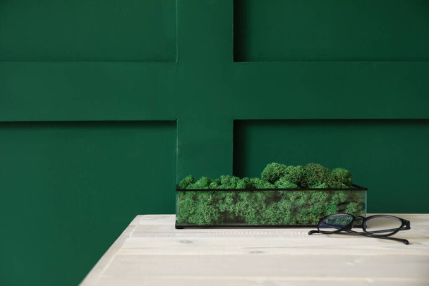 緑の壁の近くのテーブルの上に装飾的な苔と眼鏡を持つコンテナ - 写真・画像