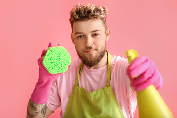 Όμορφος νεαρός άνδρας με σφουγγάρι καθαρισμού και απορρυπαντικό σε ροζ φόντο - Φωτογραφία, εικόνα