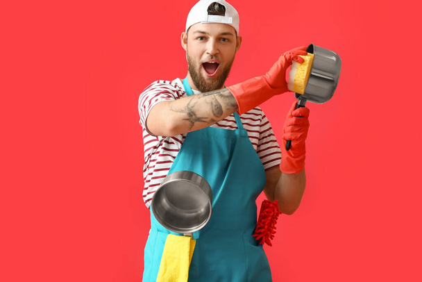 Handsome young man with sponge cleaning cooking pot on red background - Foto, afbeelding