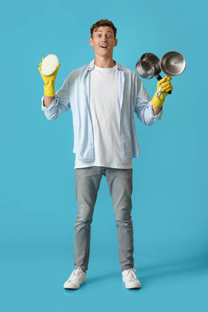 Surprised young man with cleaning sponge and cooking pots on blue background - Foto, Imagen