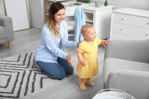 Cute baby girl in yellow dress learning to walk with mother near sofa at home - Foto, afbeelding