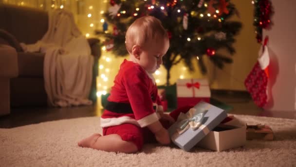 Cute baby playing with Christmas gift box under decorated Christmas tree. Families and children celebrating winter holidays. - Footage, Video