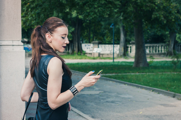 Pretty goth girl using phone in a city park - Фото, зображення