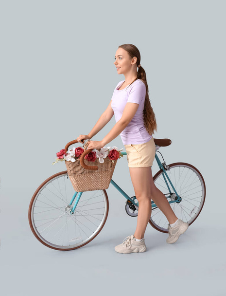 Pretty teenage girl with bicycle and flowers in bag on light background - Foto, Imagen