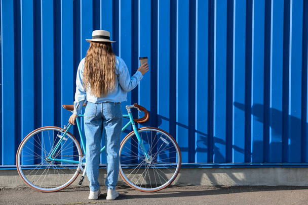 Hübsches Teenager-Mädchen mit Tasse Kaffee und Fahrrad in der Nähe von blauem Zaun - Foto, Bild