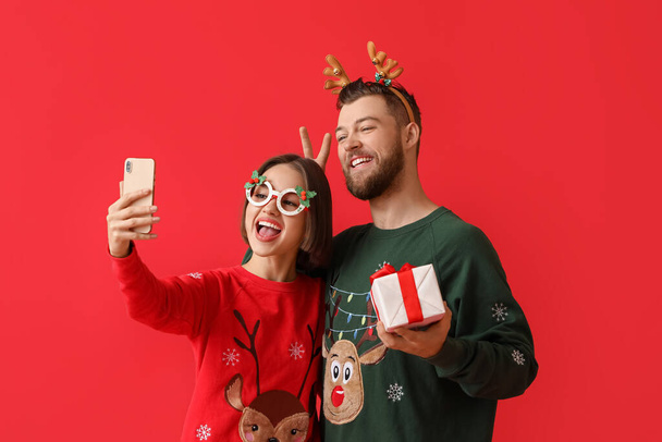 Happy young couple in stylish Christmas clothes and with gift taking selfie on color background - Foto, afbeelding