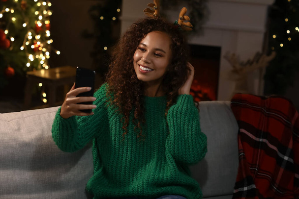 Joven mujer afroamericana en cuernos de ciervo tomando selfie en casa en la víspera de Navidad - Foto, Imagen