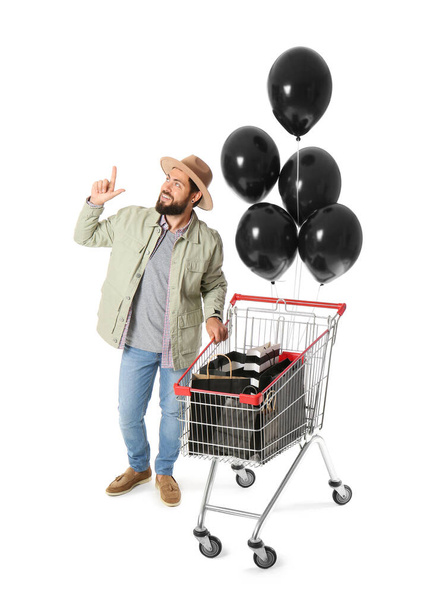Hombre guapo con bolsas de compras Viernes Negro en el carro y globos sobre fondo blanco - Foto, Imagen