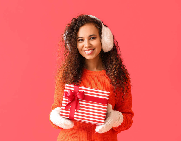 Νεαρή Αφρο-Αμερικανίδα γυναίκα σε ζεστά ακουστικά με χριστουγεννιάτικο δώρο σε κόκκινο φόντο - Φωτογραφία, εικόνα