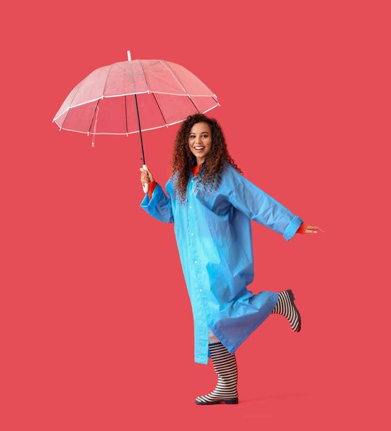 Young African-American woman wearing blue raincoat and gumboots with umbrella on red background - Foto, Imagen