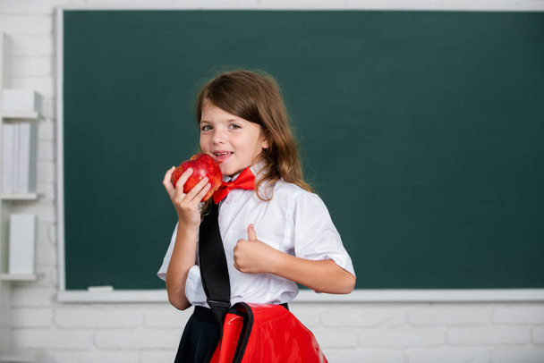 Ritratto di attraente ragazza allegra mangiare mela rossa in classe a scuola elementare. Educazione e conoscenza dei bambini. Studenti bambini. - Foto, immagini