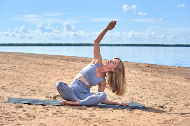 A woman does yoga on a sandy beach against the background of a river - Photo, Image