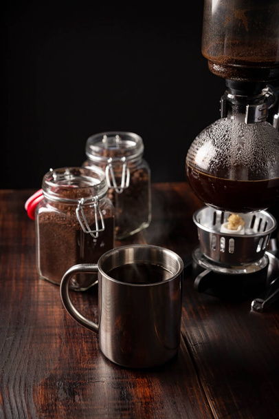Metallic cup and vacuum coffee maker also known as vac pot, siphon or syphon coffee maker and coffee glass containers on rustic wooden table - Foto, Bild