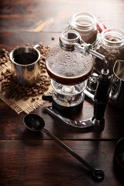 Vacuum coffee maker also known as vac pot, siphon or syphon coffee maker. Metallic cup and toasted coffee beans on rustic wooden table - Foto, Bild