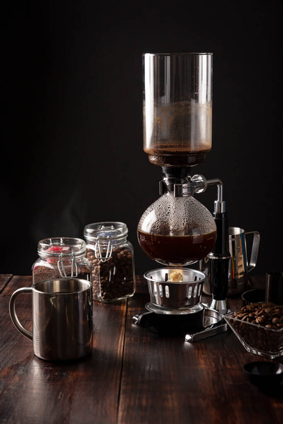 Vacuum coffee maker also known as vac pot, siphon or syphon coffee maker. Metallic cup and toasted coffee beans on rustic wooden table - Фото, зображення