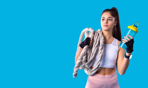 Indoor shot of attractive young active brunette young girl with ponytail hairstyle holding bottle of water and keeping the fitness rope on her shoulder. - Фото, зображення