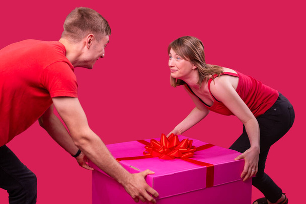 Lovely two persons man and woman wearing red t-shirts and black jeans both holding the big pink present box standing opposite and looking at each other. - Fotografie, Obrázek