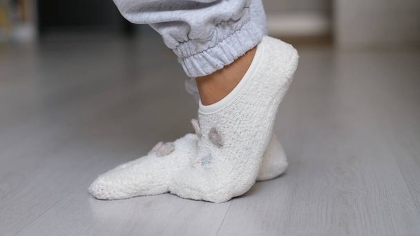 Beine einer Frau in weißen Socken, die auf dem Holzboden ihres Hauses läuft, im Hintergrund ein Sofa. Füße tragen weiße Socken auf grauem Holzboden - Foto, Bild