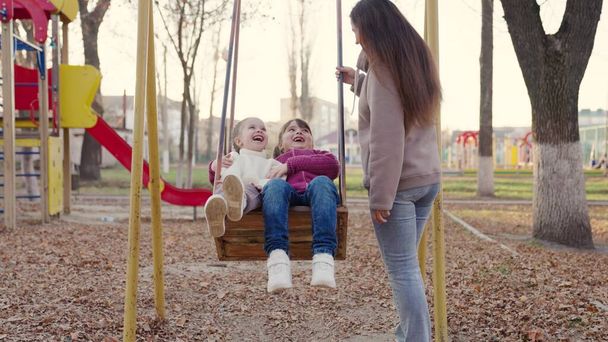 Счастливая молодая мать и маленькие дочери качаются на качелях в парке осенью. Красивые маленькие девочки сидят на качелях и смеются. Мам, дети играют вместе на детской площадке. Концепция счастливой семьи - Фото, изображение