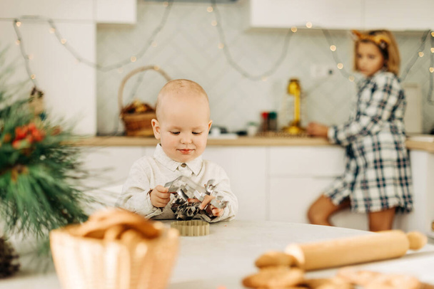 Ένα μικρό αγόρι επιλέγει κόφτες μπισκότων σε ένα τραπέζι διακοσμημένο με κλαδιά πεύκου. Γλυκά χειροποίητα δώρα για τα Χριστούγεννα. Προετοιμασία για τις γιορτές, την παραμονή Χριστουγέννων ή την παραμονή Πρωτοχρονιάς - Φωτογραφία, εικόνα