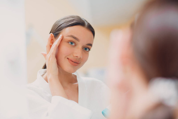 Jeune femme appliquant de la crème blanche cosmétique sur son visage au miroir - Photo, image
