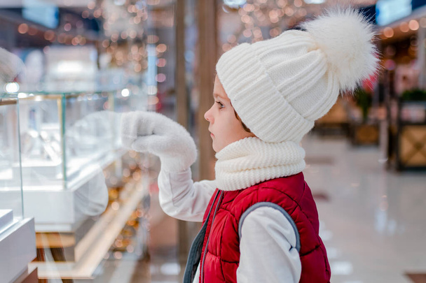 Jongens in witte pluizige hoed kijkt in de winkel door het raam en wijst iets ih showcase in de markt. - Foto, afbeelding
