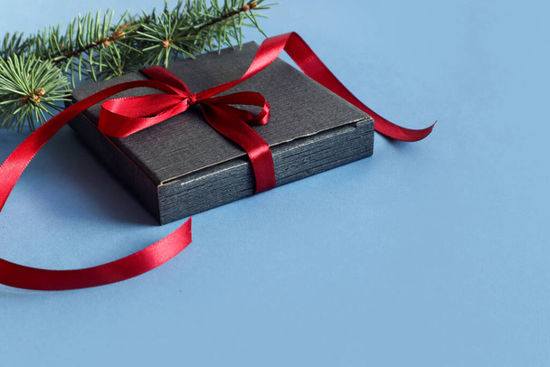 Preparing gifts for the New Year holidays: a gray gift box tied with a red ribbon, a spruce branch on a blue background, a side view, a place for text - Foto, Bild