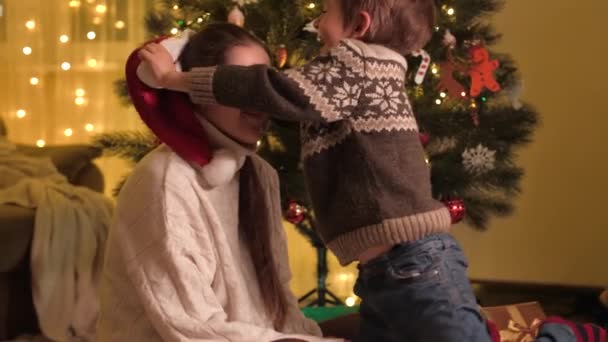 Χαρούμενο χαμογελαστό αγόρι που διασκεδάζει και βάζει το καπέλο του Άη Βασίλη στη μητέρα δίπλα στο χριστουγεννιάτικο δέντρο - Πλάνα, βίντεο