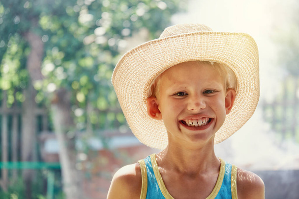 Cheerful little boy wearing large straw hat and blue sleeveless shirt posing for camera and smiles all over his mouth in sunny summer city park close view. - Foto, Bild