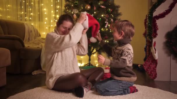 Gelukkige lachende moeder met zoon die thuis kerst viert en kerstmuts op doet. Gezinnen en kinderen vieren wintervakantie. - Video