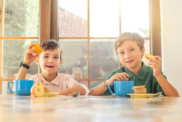 Deux heureux enfants aux yeux verts mangeant des fruits et des biscuits pour le petit déjeuner à la maison. Concept de saine alimentation pour les enfants - Photo, image