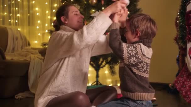 Jonge moeder zet Santa hoed op haar zoon en knuffelt hem onder de kerstboom. Gezinnen en kinderen vieren wintervakantie. - Video