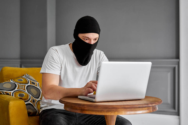 Portret nierozpoznawalnego młodego hakera w czarnej masce przy użyciu laptopa. Pojęcie bezpieczeństwa cybernetycznego i ochrony danych - Zdjęcie, obraz
