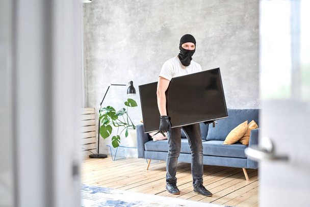 Modern pahalı televizyonu çalan siyah kar maskeli bir hırsız. Maskeli yüz. Hırsız evden televizyon çalıyor. - Fotoğraf, Görsel
