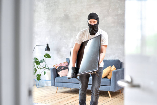 Dieb mit schwarzer Sturmhaube stiehlt modernen teuren Fernseher Maskiertes Gesicht. Mann klaut Fernseher aus Haus - Foto, Bild