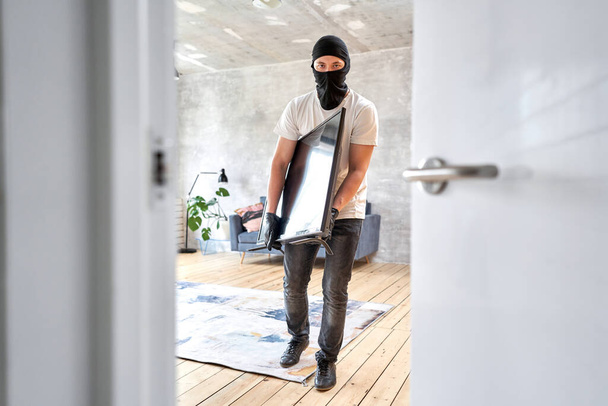 Thief with black balaclava stealing modern expensive television. Masked face. Man burglar stealing tv set from house - Fotografie, Obrázek