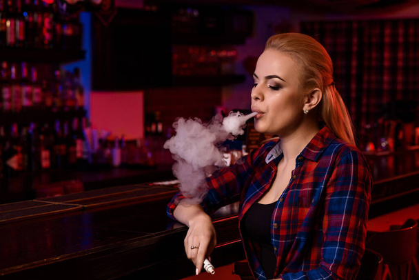 Young pretty woman smoke an electronic cigarette at the vape bar - Фото, изображение