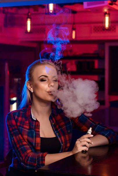 Νεαρή όμορφη γυναίκα σε ένα πουκάμισο σε ένα κλουβί καπνίζει ένα ηλεκτρονικό τσιγάρο στο μπαρ - Φωτογραφία, εικόνα