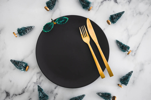 golden cutlery on top of black dining plate with Chrismtas decorations all around on marble table, concept of festive season and holiday meals - Foto, Imagen