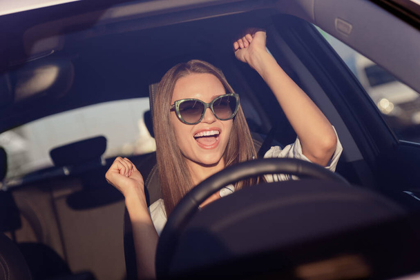 Photo of sweet lucky young lady dressed white shirt dark eyewear smiling rising fists driving automobile outdoors urban route. - Foto, Imagen