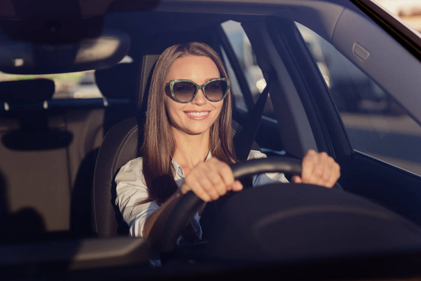 Photo of charming sweet young lady dressed white shirt dark eyewear smiling driving automobile outdoors urban route. - Foto, imagen