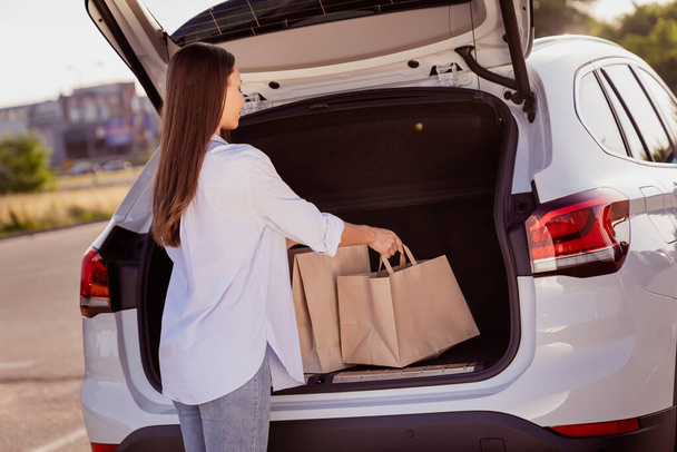 Фото очаровательной красивой молодой женщины носить белую рубашку положить сумки багажник автомобиля возле улицы города. - Фото, изображение
