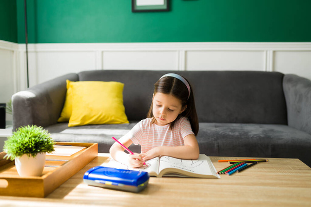 Das gehorsame kleine Mädchen macht seine Hausaufgaben allein im Wohnzimmer. Grundschüler spielen mit Malbuch  - Foto, Bild
