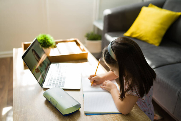 Virtuális oktatási koncepció. Okos óvodás srác írja a házi feladatát egy jegyzetfüzetbe, miközben nézi az online tanárát egy videóhíváson.  - Fotó, kép
