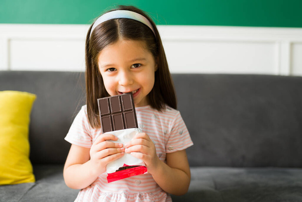 Çikolataya bayılırım. Pembe elbiseli beyaz kız koltukta dinlenirken çikolata yiyor.  - Fotoğraf, Görsel