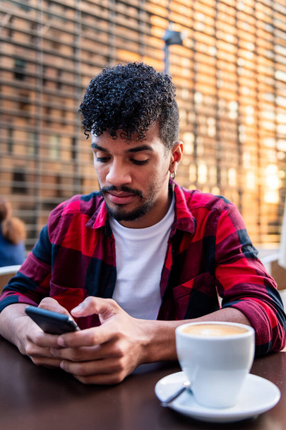 vertical photo of a young latin man sitting on a terrace with a coffee and looking at his phone, concept of technology and urban lifestyle - Zdjęcie, obraz