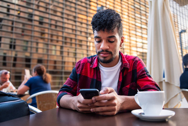 young latin man consulting his phone while sitting with a coffee on the terrace of a coffee shop, concept of technology and urban lifestyle - Zdjęcie, obraz